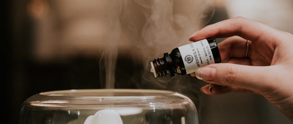 A hand holds an essential oil bottle over a diffuser releasing vapor
