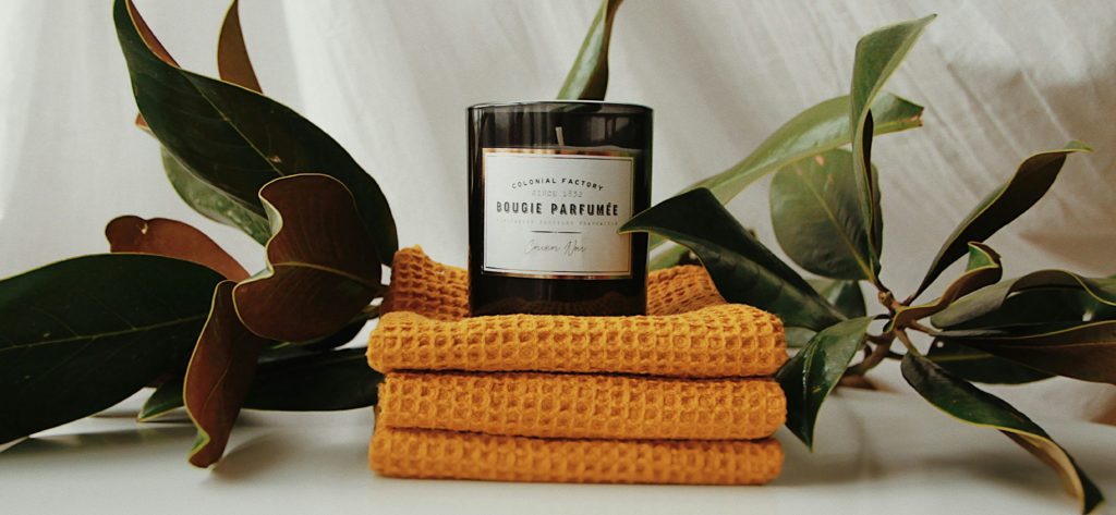 A black candle labeled "Bougie Parfumée" sits on top of folded yellow fabric, surrounded by green leaves against a white background.