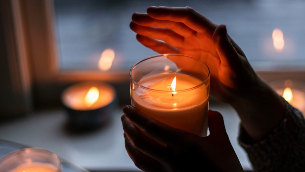 Two hands hold a lit candle in a glass container, with several other candles burning in the background.