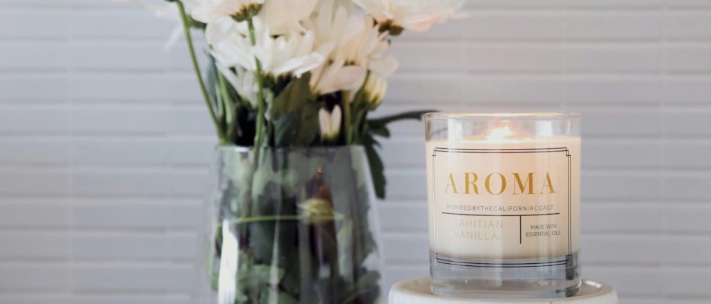 A lit candle in a glass jar labeled "Aroma," next to a clear vase filled with white flowers, set against a light-colored, tiled background.