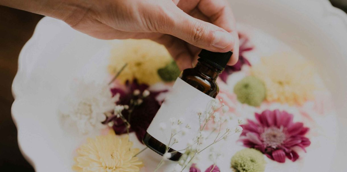 Hand holding a small glass bottle of essential oil, against a background of various colorful flowers.