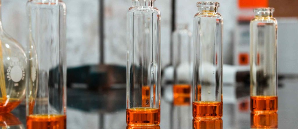 Several tall, clear glass vials containing a small amount of orange essential oil, arranged on a reflective surface in a laboratory setting