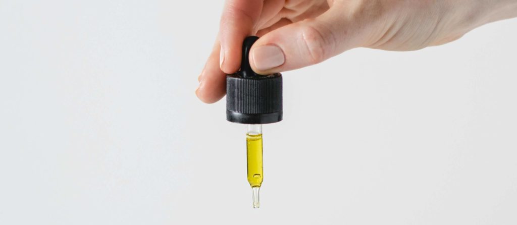 Hand holding a black-capped dropper with a yellow essential oil inside, against a plain white background