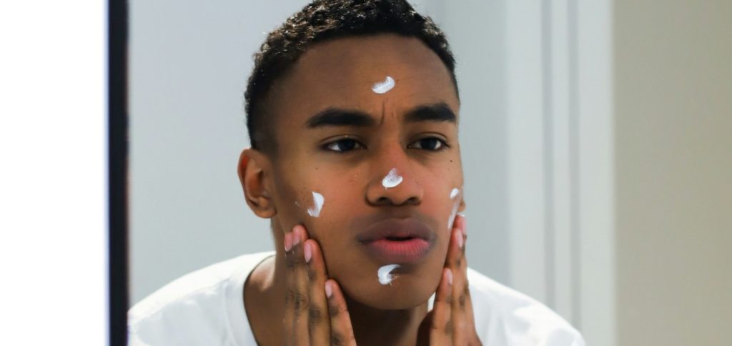 A young man is looking at himself in a mirror while applying white cream or lotion to his face