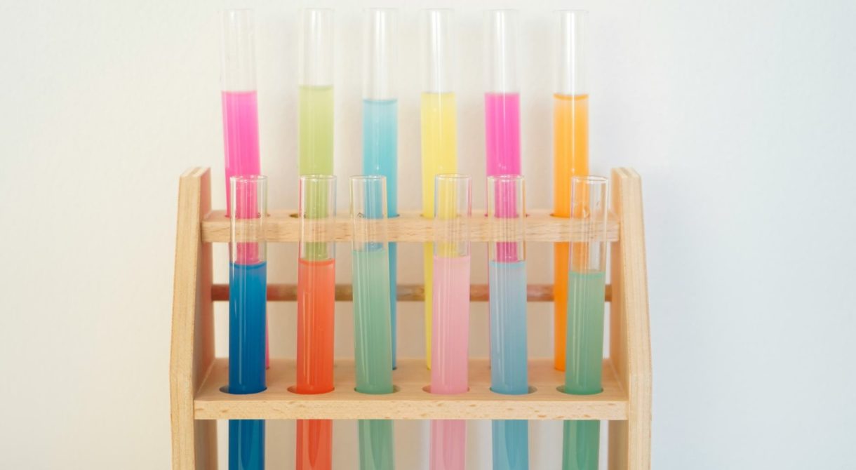 A wooden rack holding several test tubes, each filled with colorful liquids in shades of pink, blue, green, orange, and yellow.