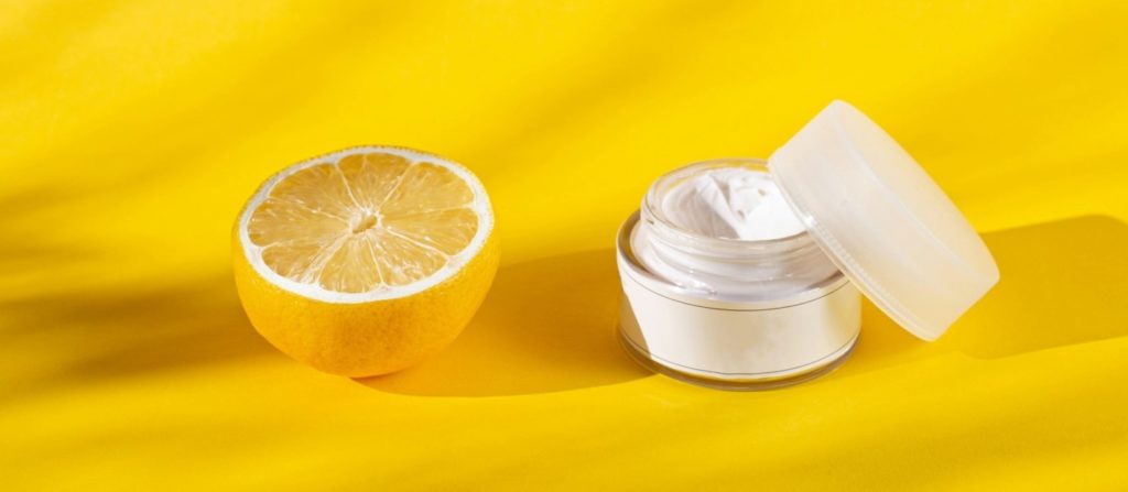 A fresh half-cut lemon placed next to an open jar of white cream on a bright yellow surface