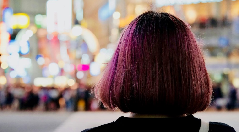 A girl with short, straight hair dyed in a gradient of purple and pink stands facing away from the camera