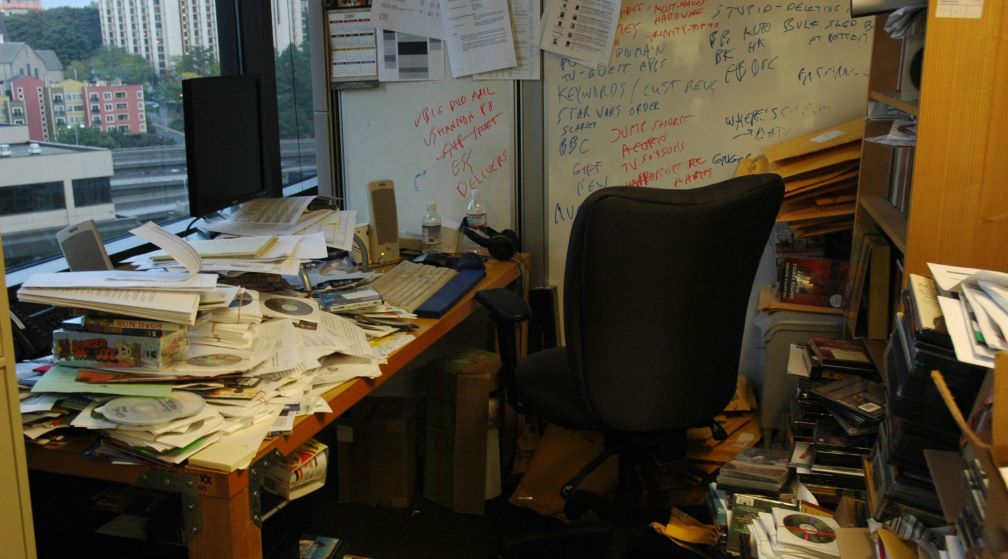 A cluttered office desk with piles of papers, documents, and CDs scattered across it.