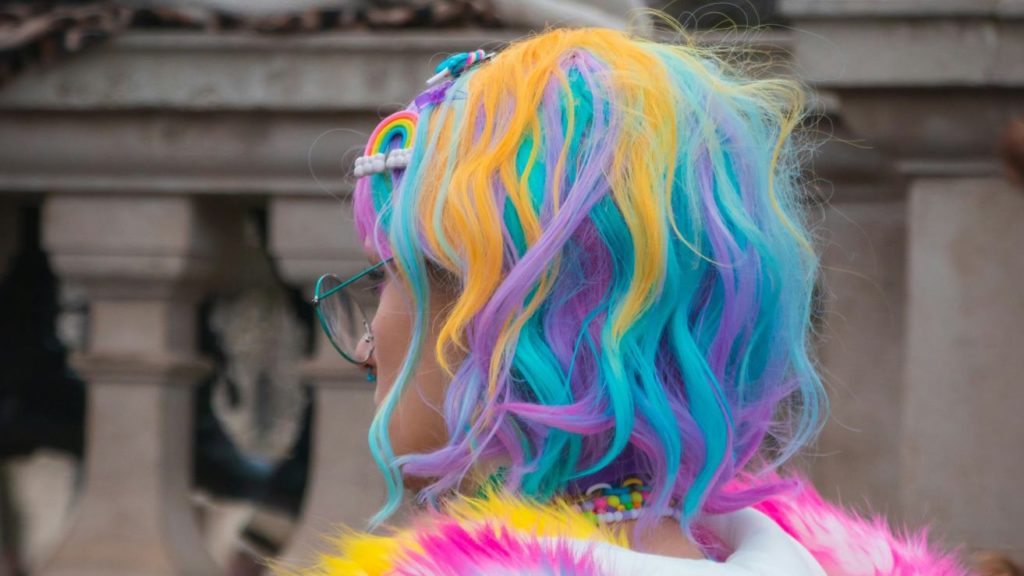 A girl with curly, shoulder-length hair dyed in bright rainbow colors, including yellow, blue, purple, and pink