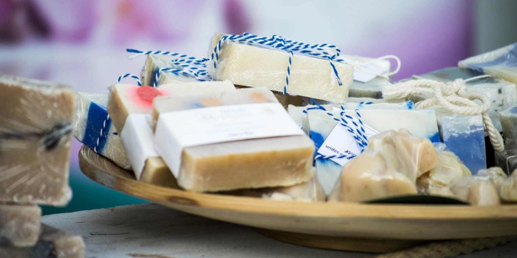 A wooden tray filled with artisan soap bars wrapped in plastic and white labels, each tied with blue and white twine. 