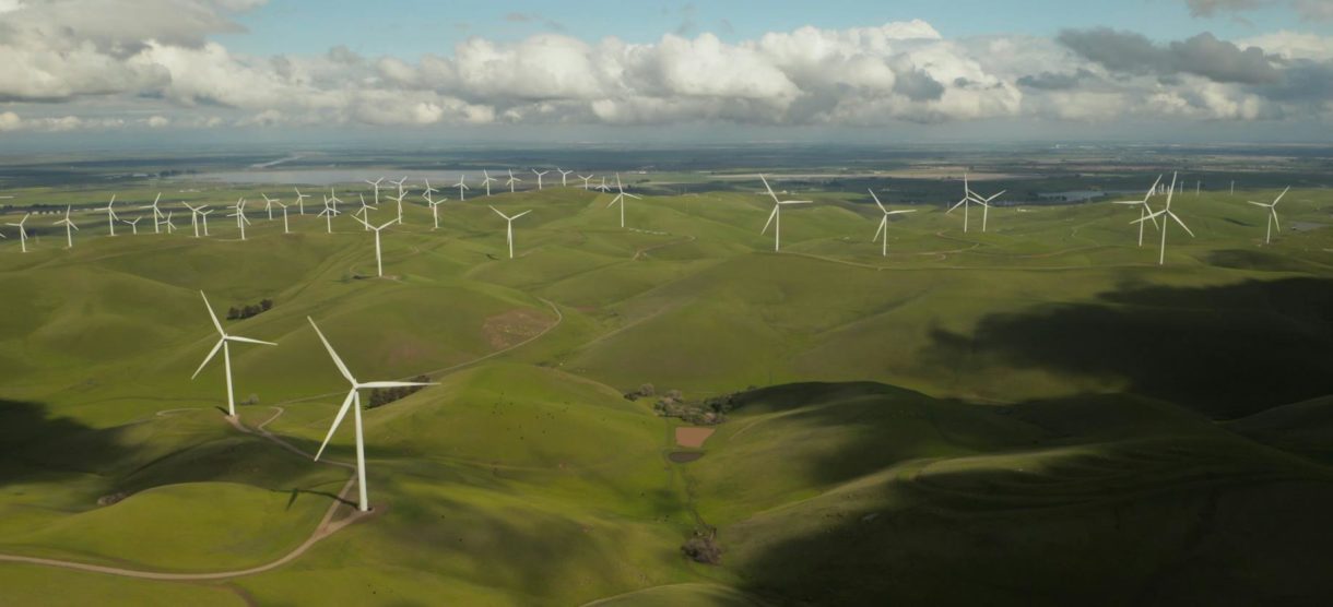 A vast green landscape dotted with wind turbines spread across rolling hills under a cloudy sky, representing a wind farm generating renewable energy