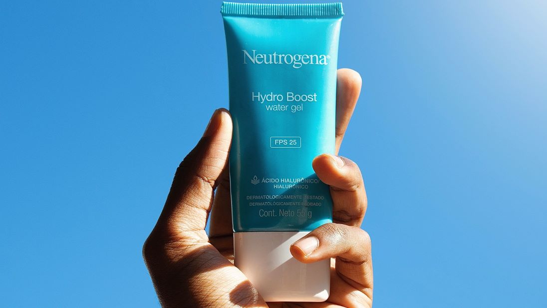 A hand holding a tube of water gel sunscreen against a blue sky.