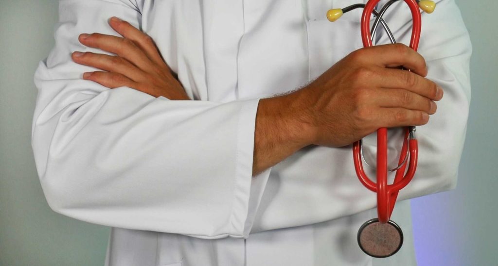 A close-up of a person wearing a white lab coat, holding a red stethoscope with arms crossed