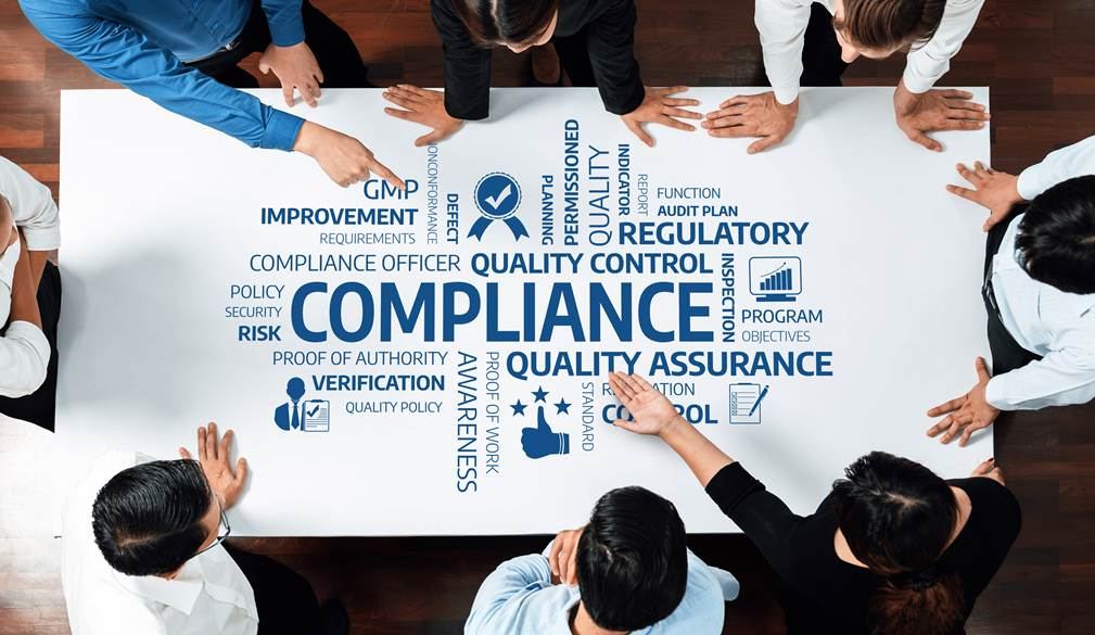 people standing and pointing at a white table top that has compliance, regulatory, quality assurance, quality control written on it